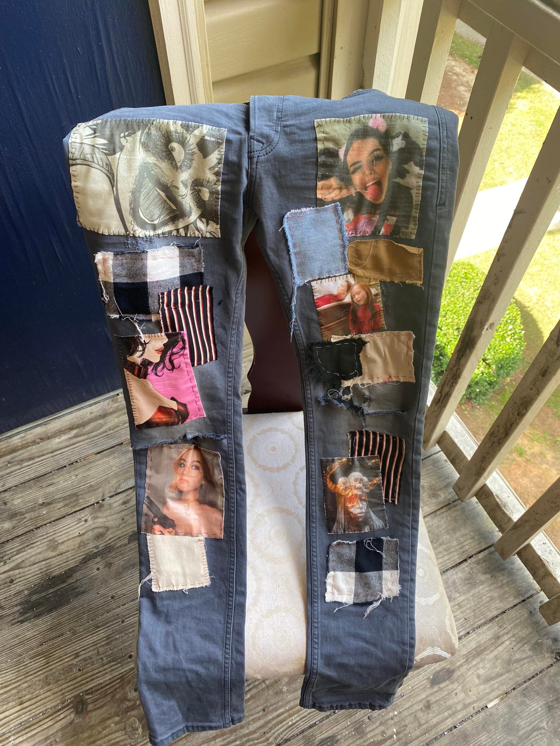 Pair of gray jeans with various colorful fabric patches and photo prints sewn onto the legs, displayed on a chair outside on a wooden porch next to a railing.