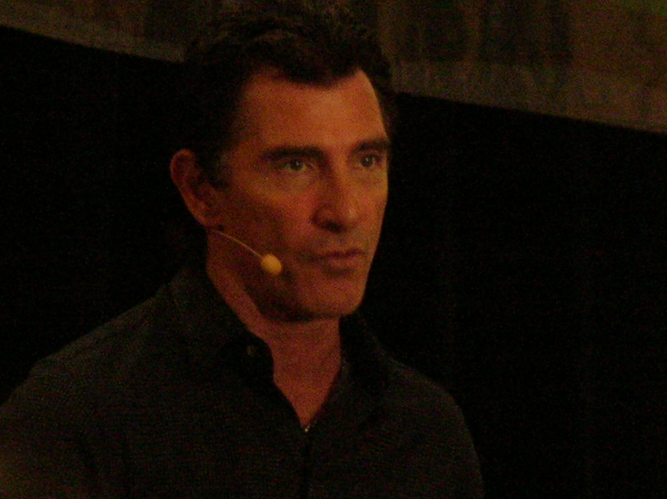 A man with dark hair wearing a headset microphone speaks against a dark background.