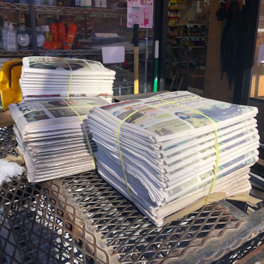 Two bundles of newspapers, tied with yellow bands, rest on a metal grate outside a store with visible shelves and products in the background. Sunlight casts shadows on the newspapers and the ground.