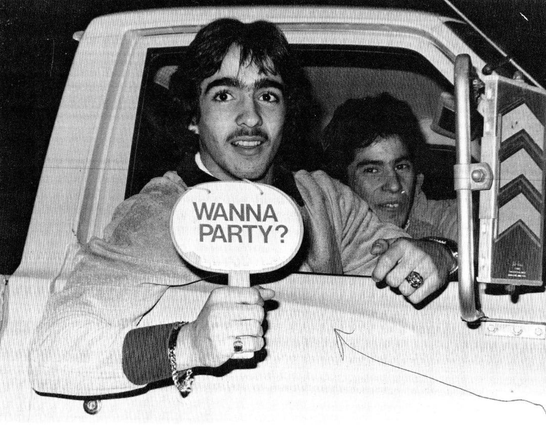 Two young men sit in a car at night. The man in front holds a sign reading “WANNA PARTY?” and looks at the camera, smiling. The other man sits behind him, partially visible, with a neutral expression.