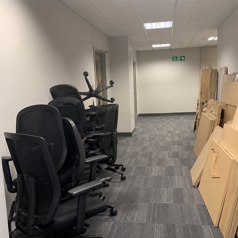 Office chairs are stacked along the left wall of a hallway, and piles of wooden boards or panels are leaning against the right wall. The hallway has gray carpet and overhead fluorescent lighting.