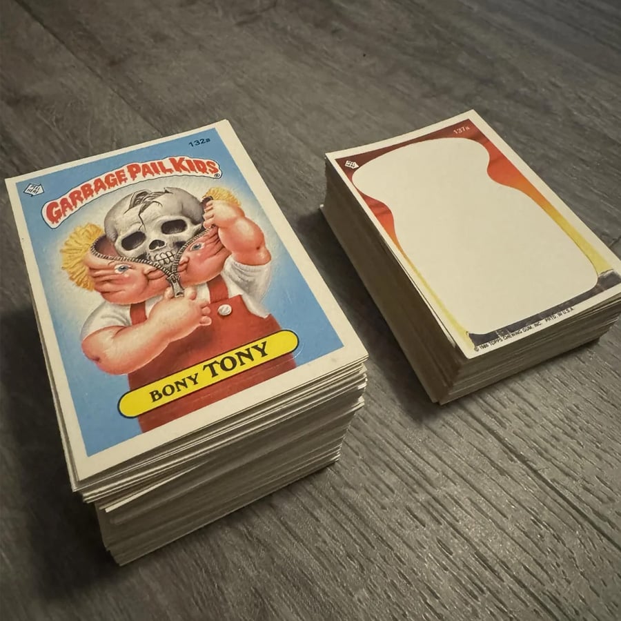 Two stacks of trading cards on a wood floor: one stack shows a Garbage Pail Kids “Bony Tony” card, the other shows cards with a blank design.