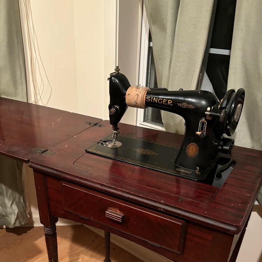 A vintage black Singer sewing machine is built into a dark wooden table, positioned near windows with light green curtains. The sewing machine has a classic design with gold detailing.