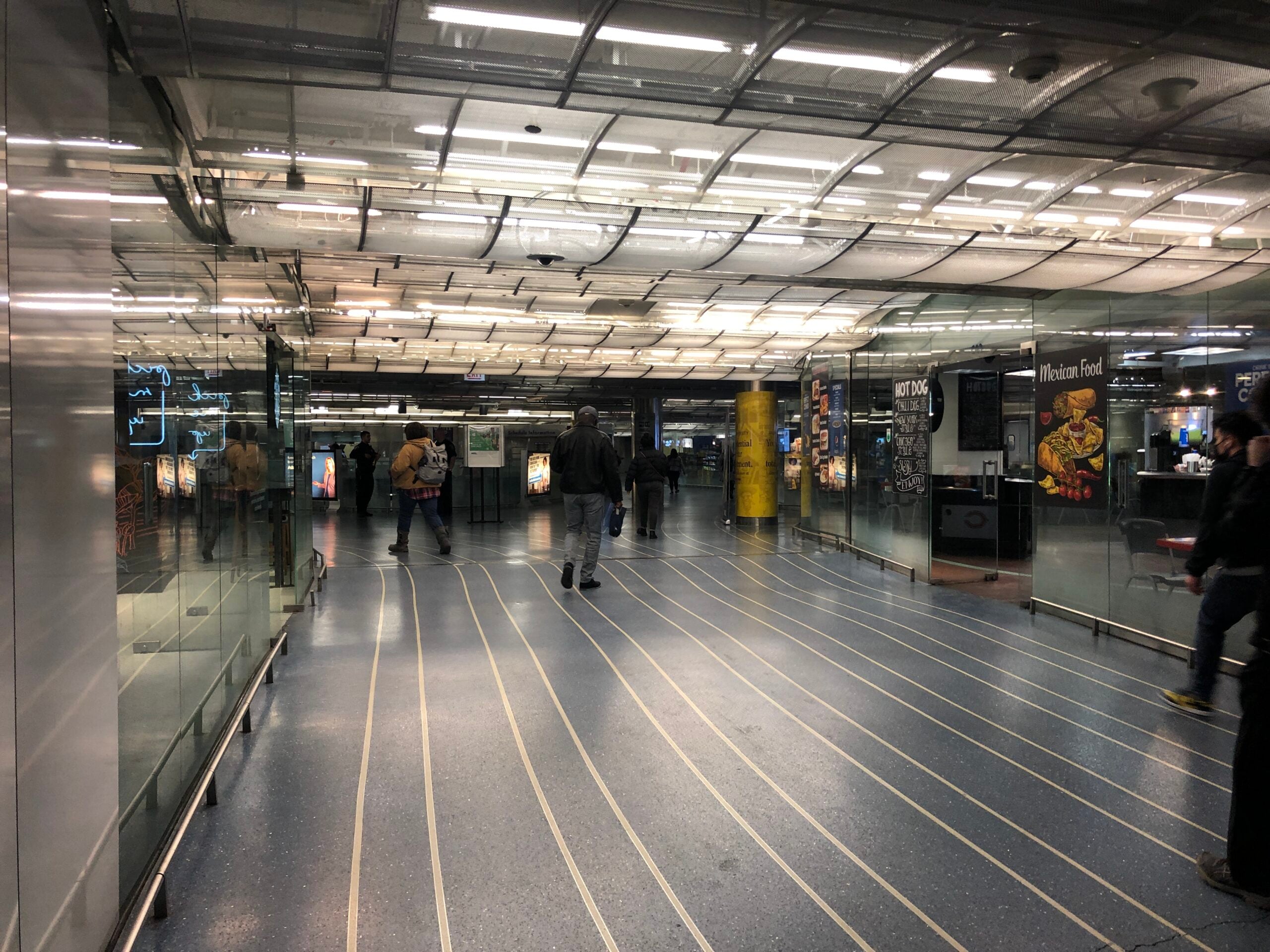 People walk through a modern, well-lit subway or train station corridor with glass walls, blue flooring with white lines, and illuminated advertisements along the sides.