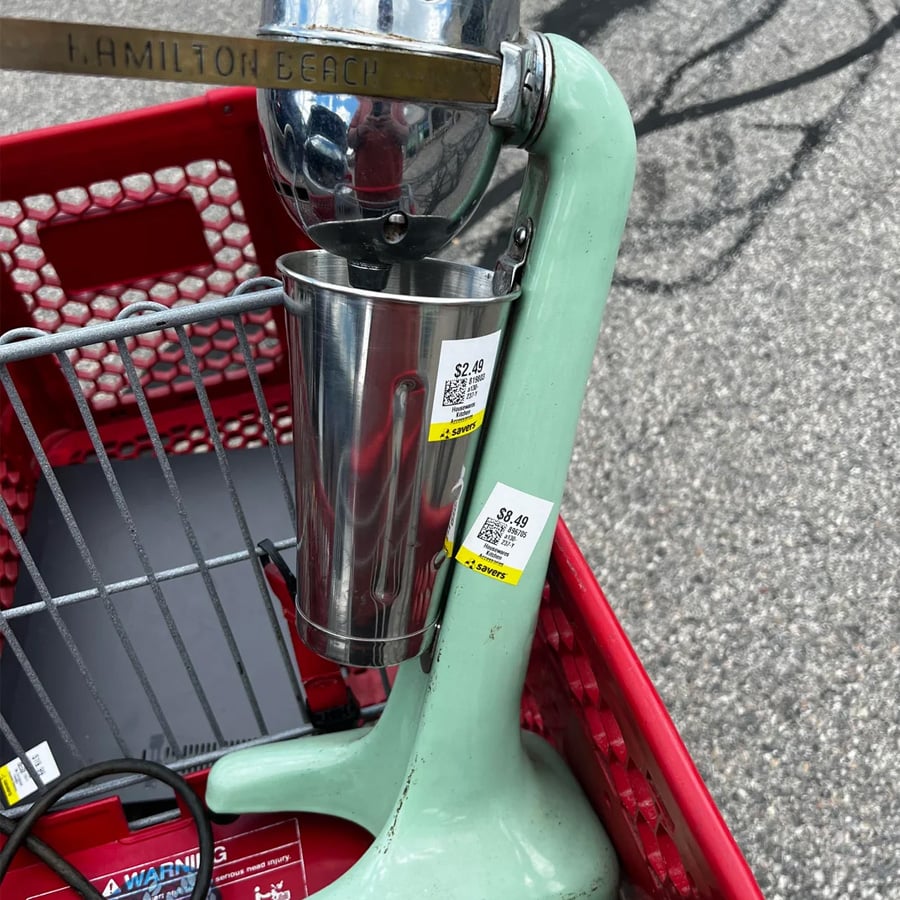 A vintage Hamilton Beach milkshake mixer sits in a red shopping cart, with price tags on its base and metal cup. The cart is outdoors on a paved surface.