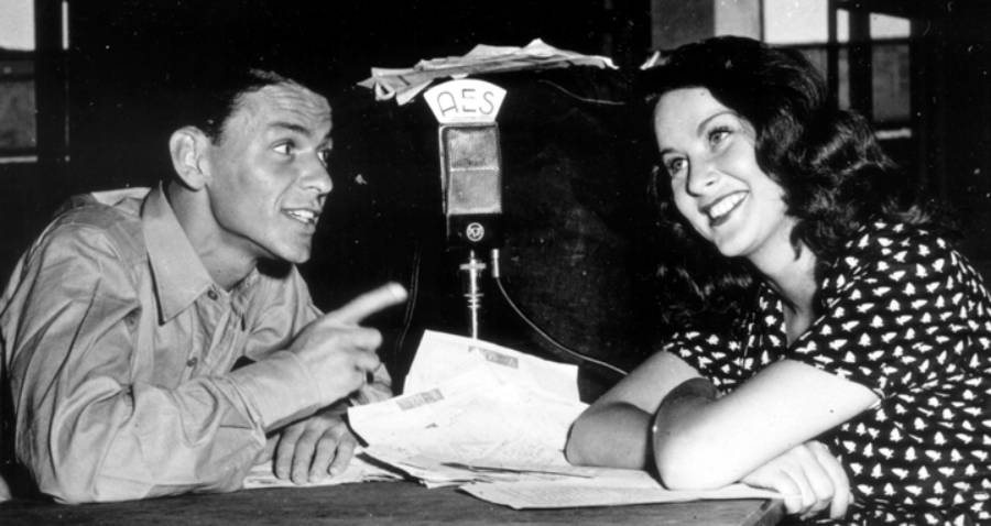 A man and woman sit at a table with papers in front of a vintage microphone labeled “AES.” The man gestures while talking, and the woman smiles, listening to him.