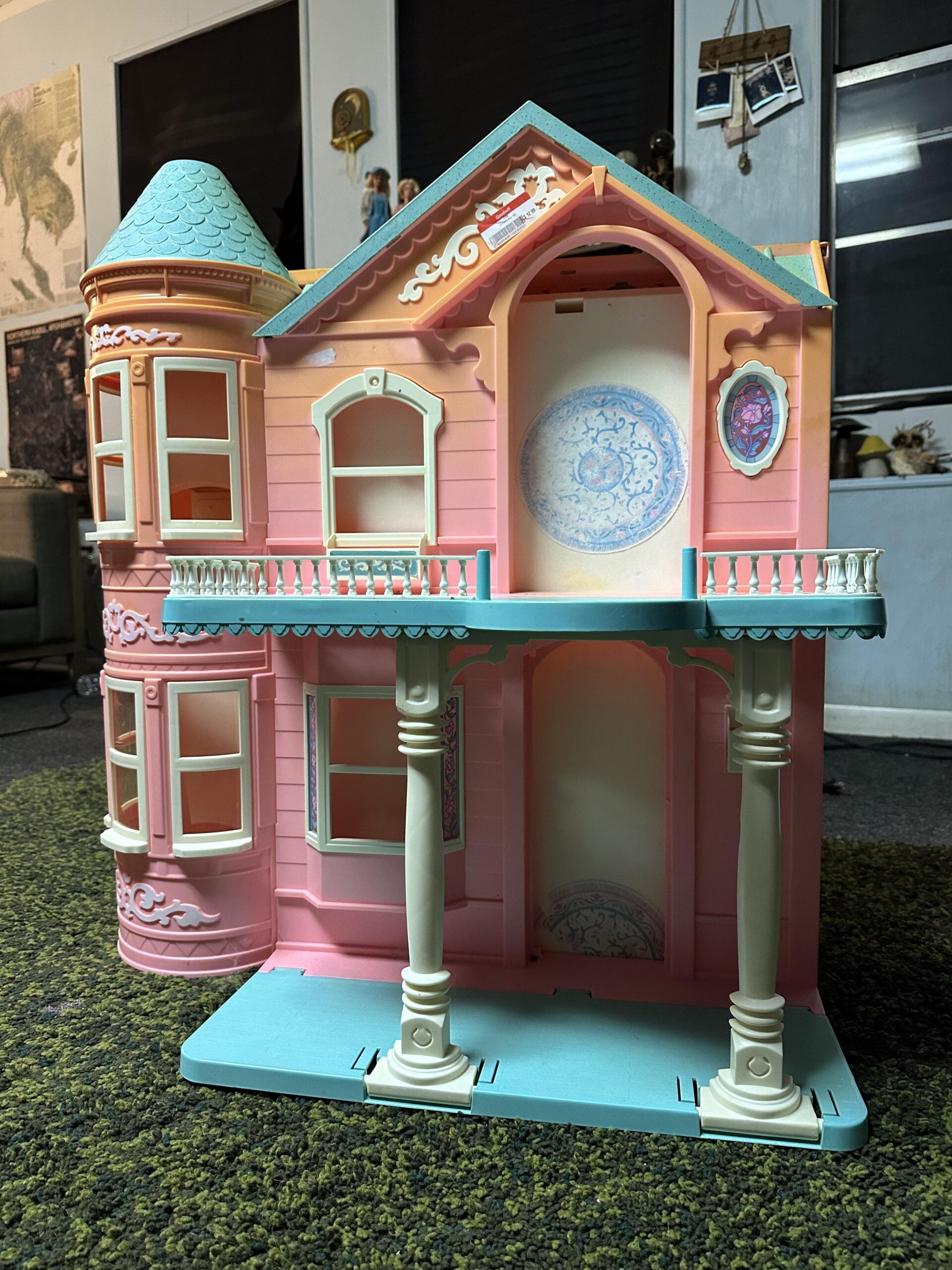 A pastel pink and blue toy dollhouse with two stories, a turret, decorative columns, arched windows, and ornate detailing, displayed indoors on a green carpet.