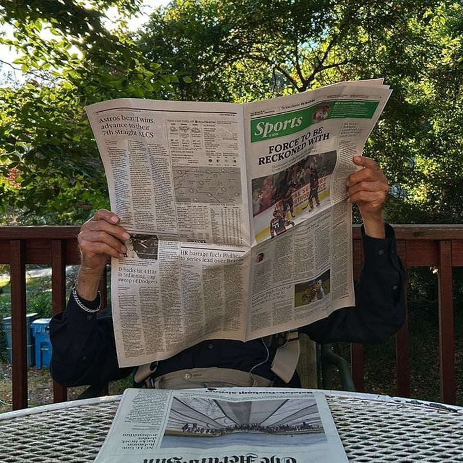 A person sits outside at a table, holding up and reading a large newspaper that covers their face. Another newspaper lies folded on the table. Sunlight and greenery are visible in the background.