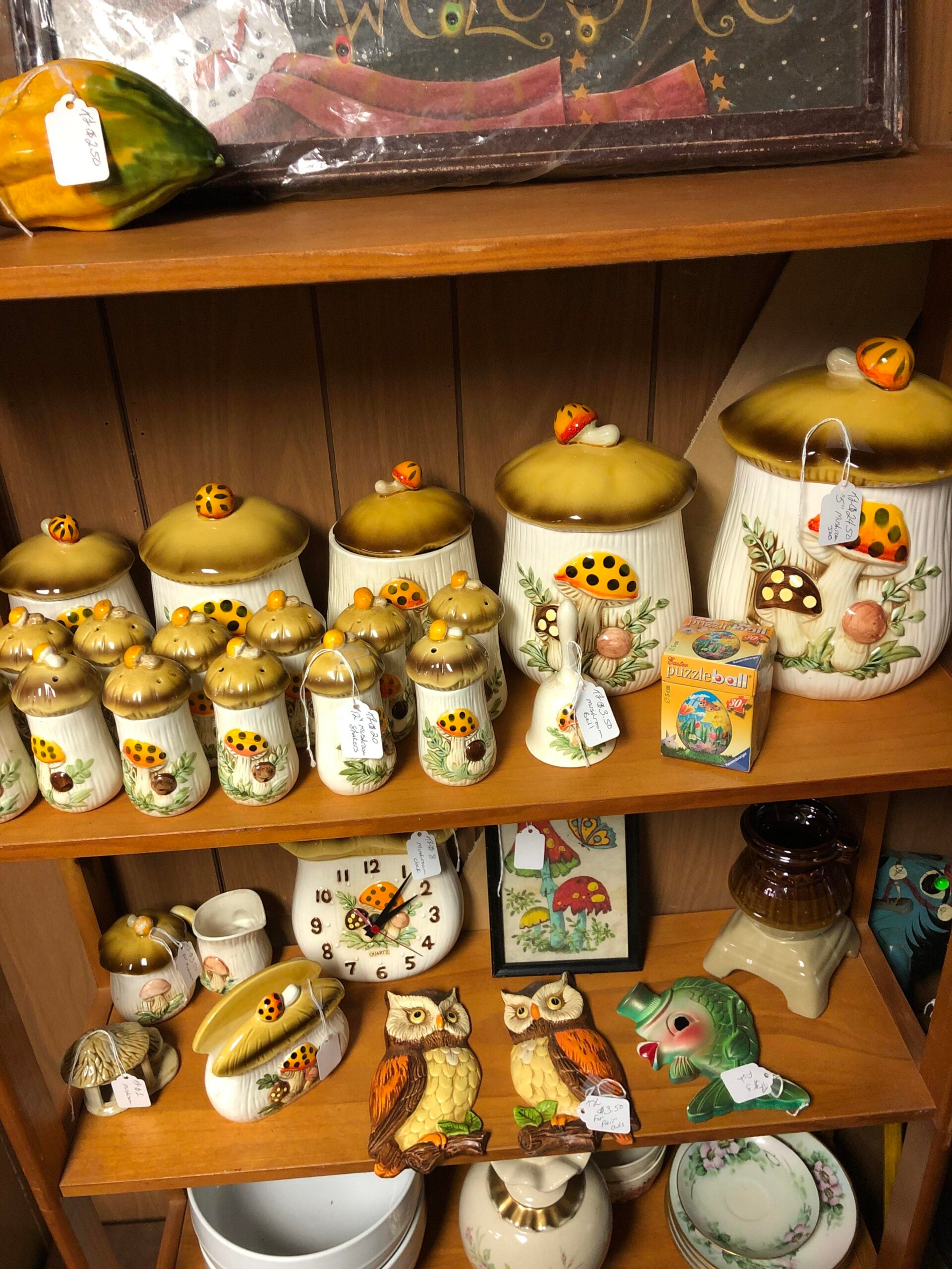 A wooden shelf holding various vintage owl figurines, mushroom-themed canisters, spice jars with yellow lids, a green fish figurine, a clock, and other assorted decorative items, all with visible price tags.