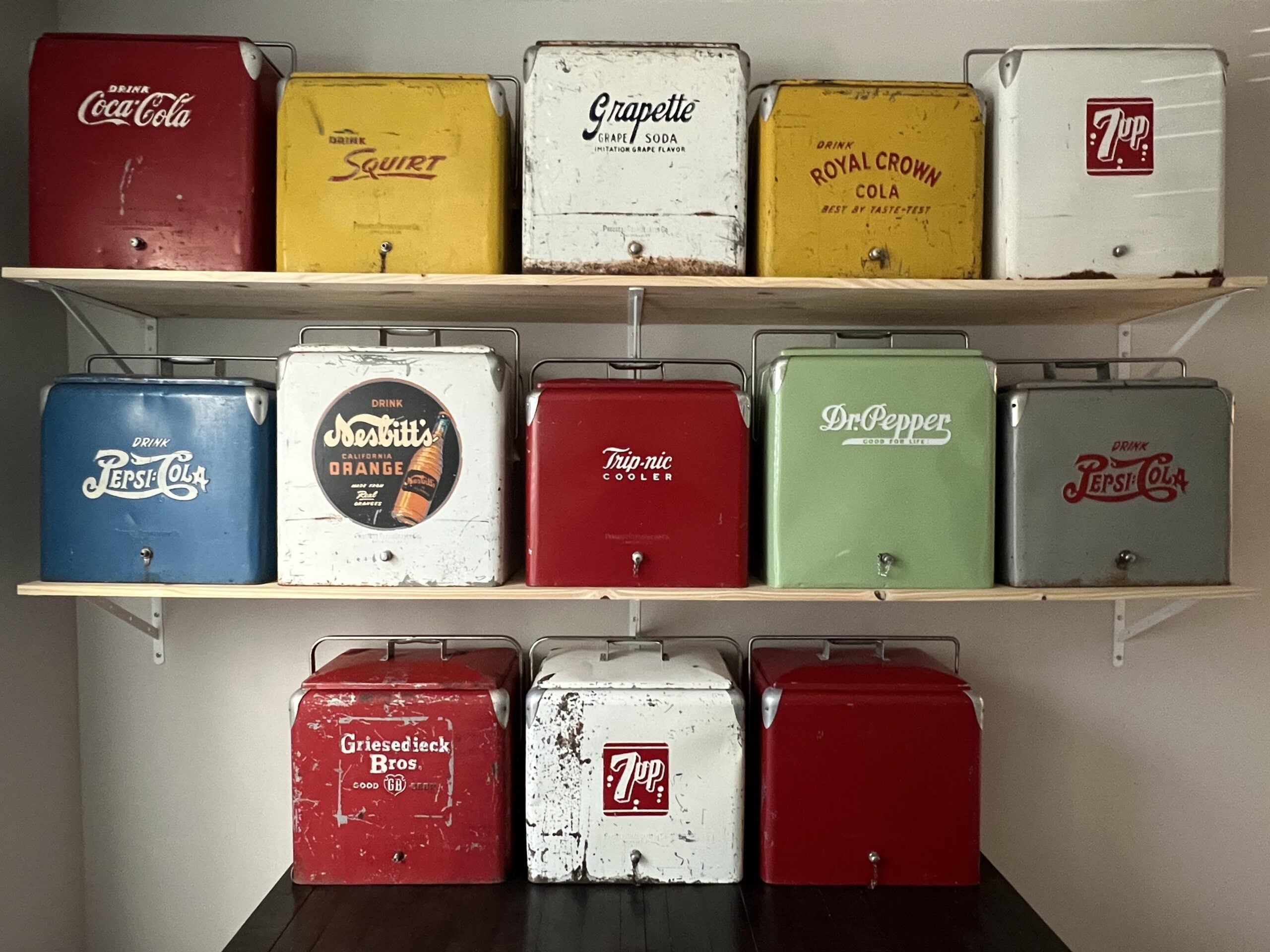 Twelve vintage metal coolers with old soda brand logos, including Coca-Cola, Squirt, Pepsi, Dr Pepper, and 7 Up, are arranged on two shelves against a wall.
