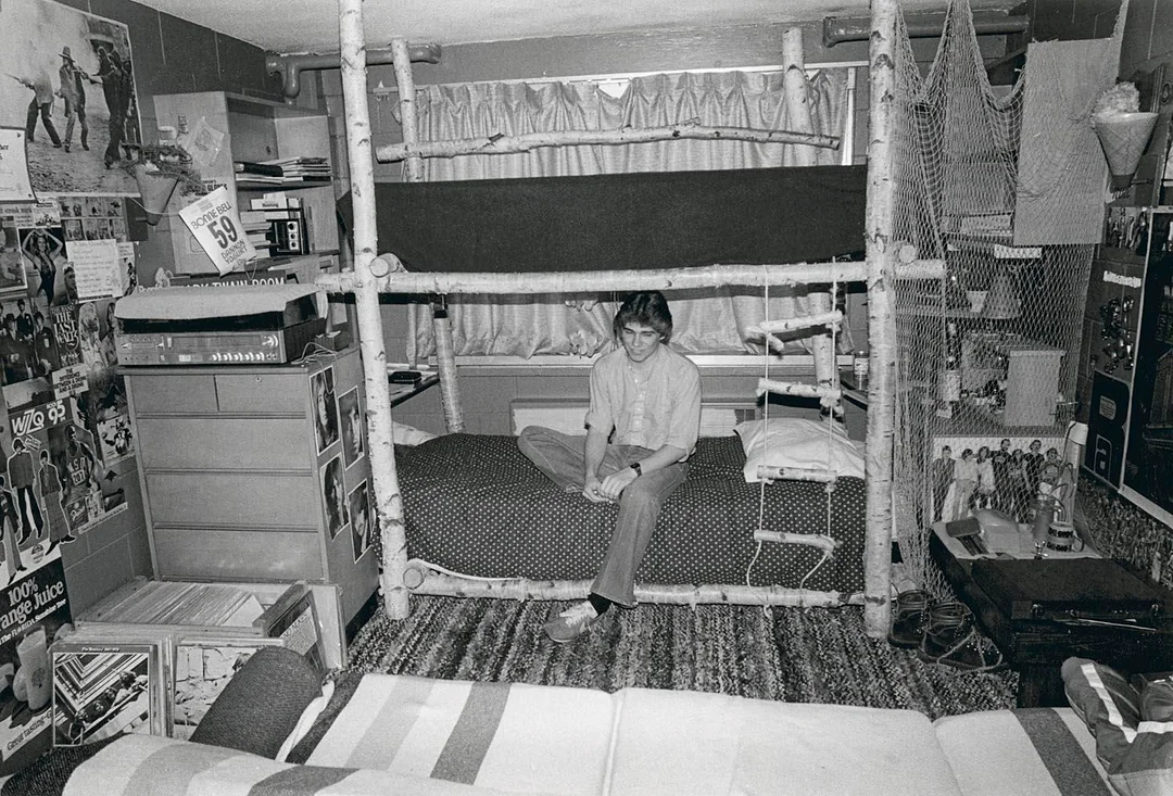 A person sits on a bunk bed in a cluttered bedroom filled with posters, books, records, and personal items. Rugs cover the floor, and shelves and storage line the walls. The room looks lived-in and personalized.