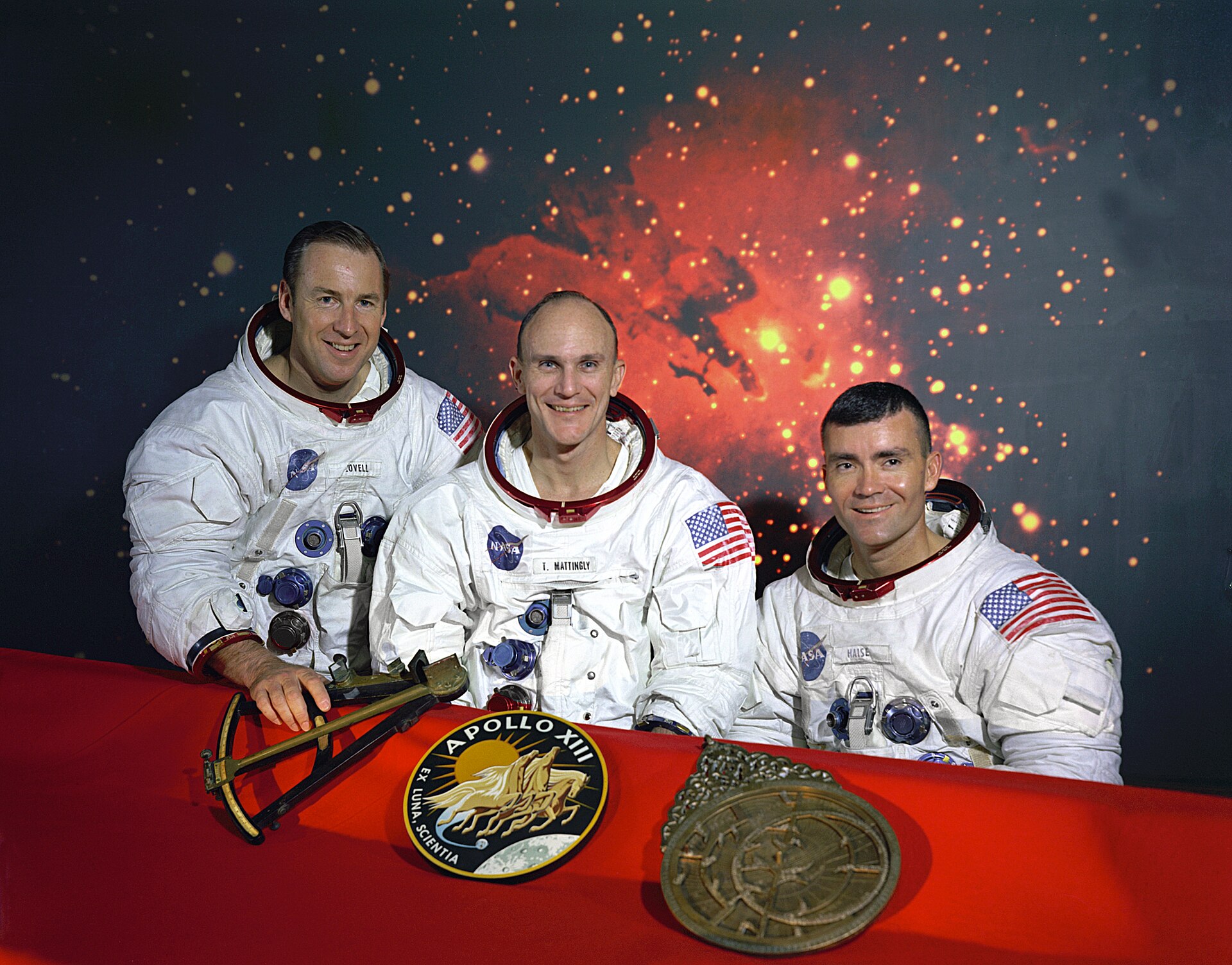 Three astronauts in white space suits pose at a red table with Apollo 13 memorabilia in front of a cosmic background. They are smiling, with mission patches and medals displayed before them.