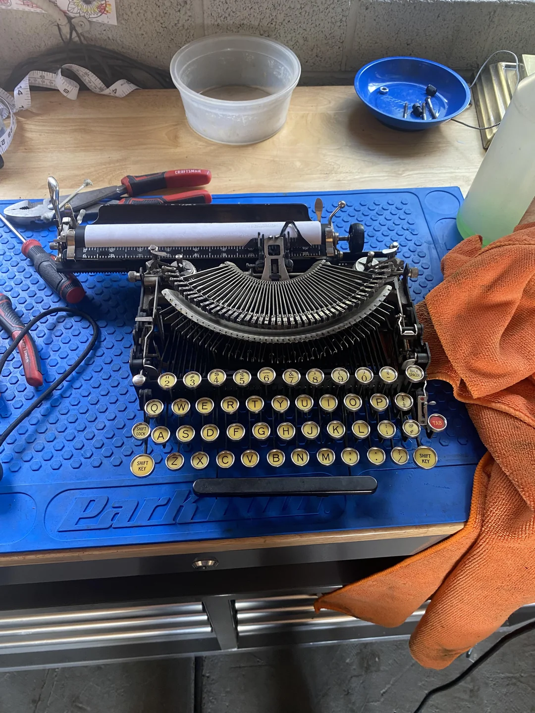 An old typewriter sits on a blue mat on a workbench, surrounded by tools, an orange cloth, and a clear plastic container.