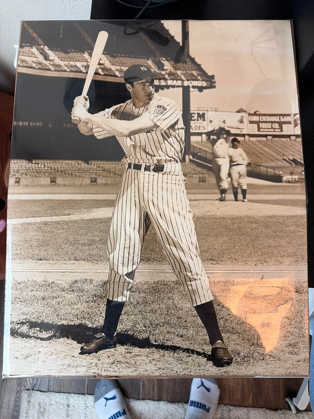 A vintage photo shows a New York Yankees baseball player in pinstripes holding a bat, posing at home plate in an empty stadium, with two other players visible in the background.