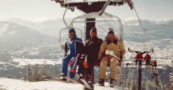 Three people in winter clothing ride a ski lift over a snowy mountain landscape, with trees below and distant mountains in the background. Other skiers are visible on lifts behind them.