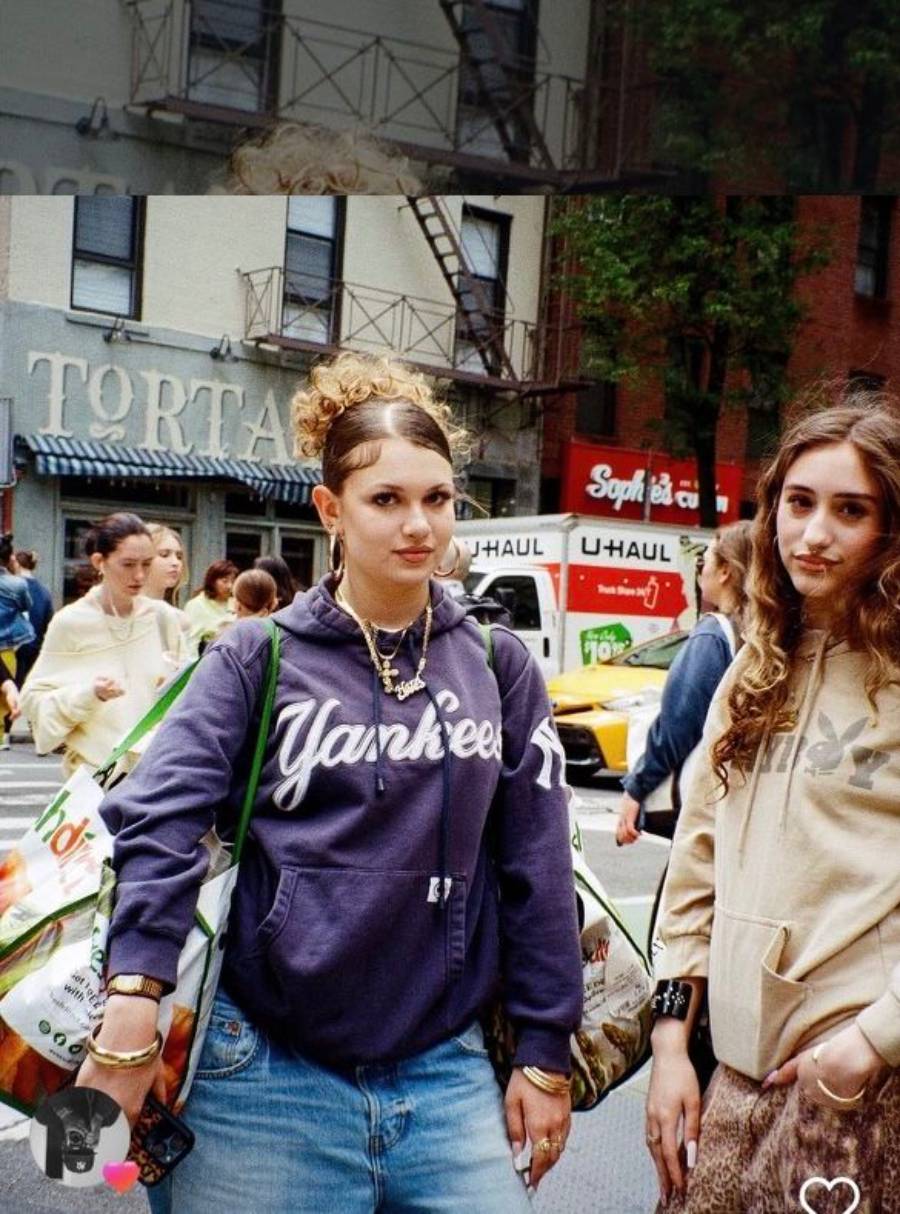 Two young women pose on a city street. One wears a Yankees hoodie and carries shopping bags; the other wears a tan hoodie. People and parked U-Haul vans are in the background near buildings.