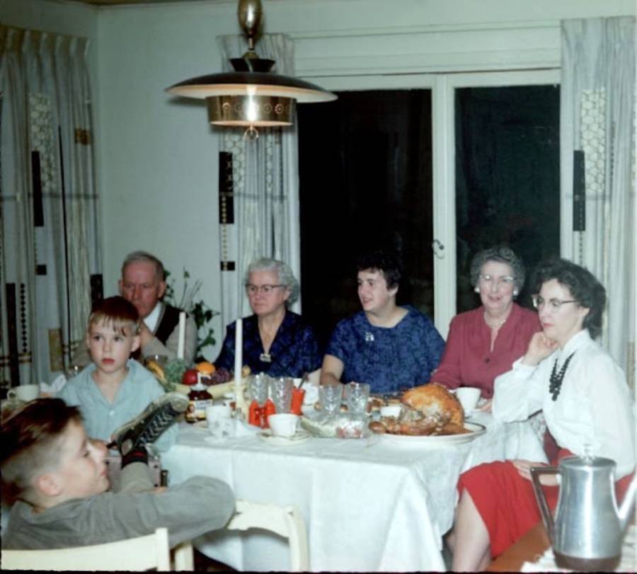 A family sits around a dining table with a turkey and dishes, appearing to enjoy a meal together. There are adults and children, and the scene has a warm, vintage feel.