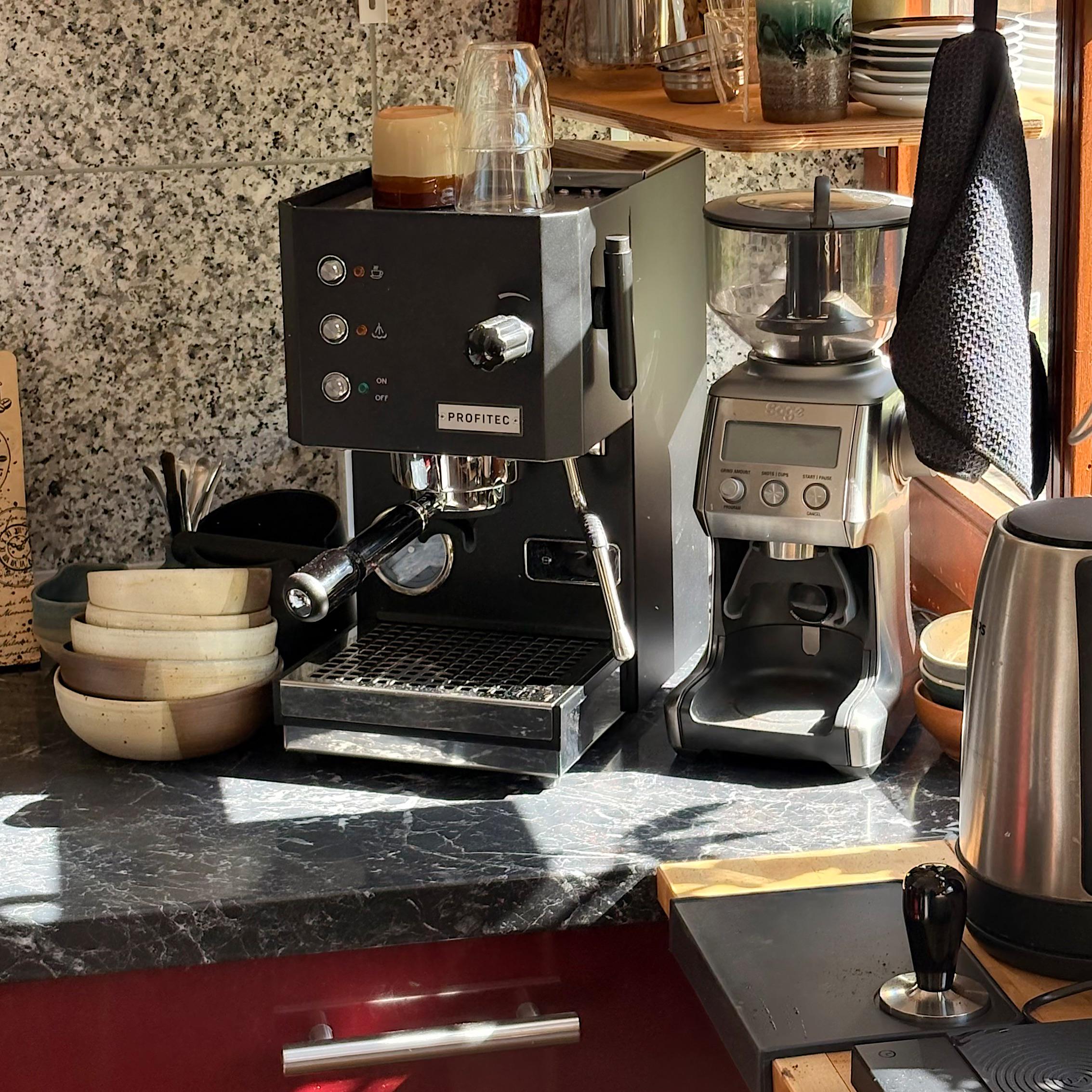 A coffee station with an espresso machine, grinder, kettle, bowls, glass cup, and tamper on a sunlit marble countertop, next to a window.