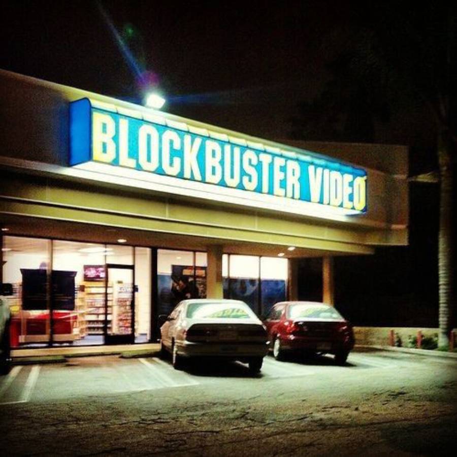 A brightly lit Blockbuster Video store at night with two cars parked in front; the blue and yellow Blockbuster sign is prominently displayed above the entrance.