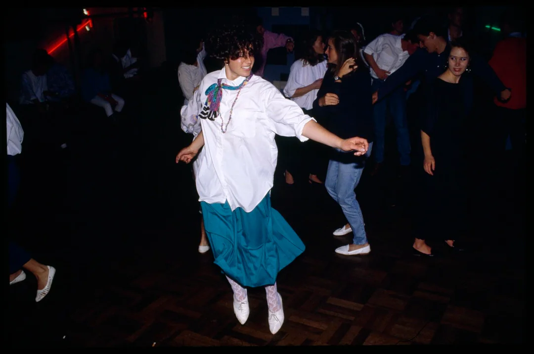 A woman in a white blouse, teal skirt, and scarf dances energetically at a dimly lit party with other people. Neon lights and wooden flooring are visible in the background.