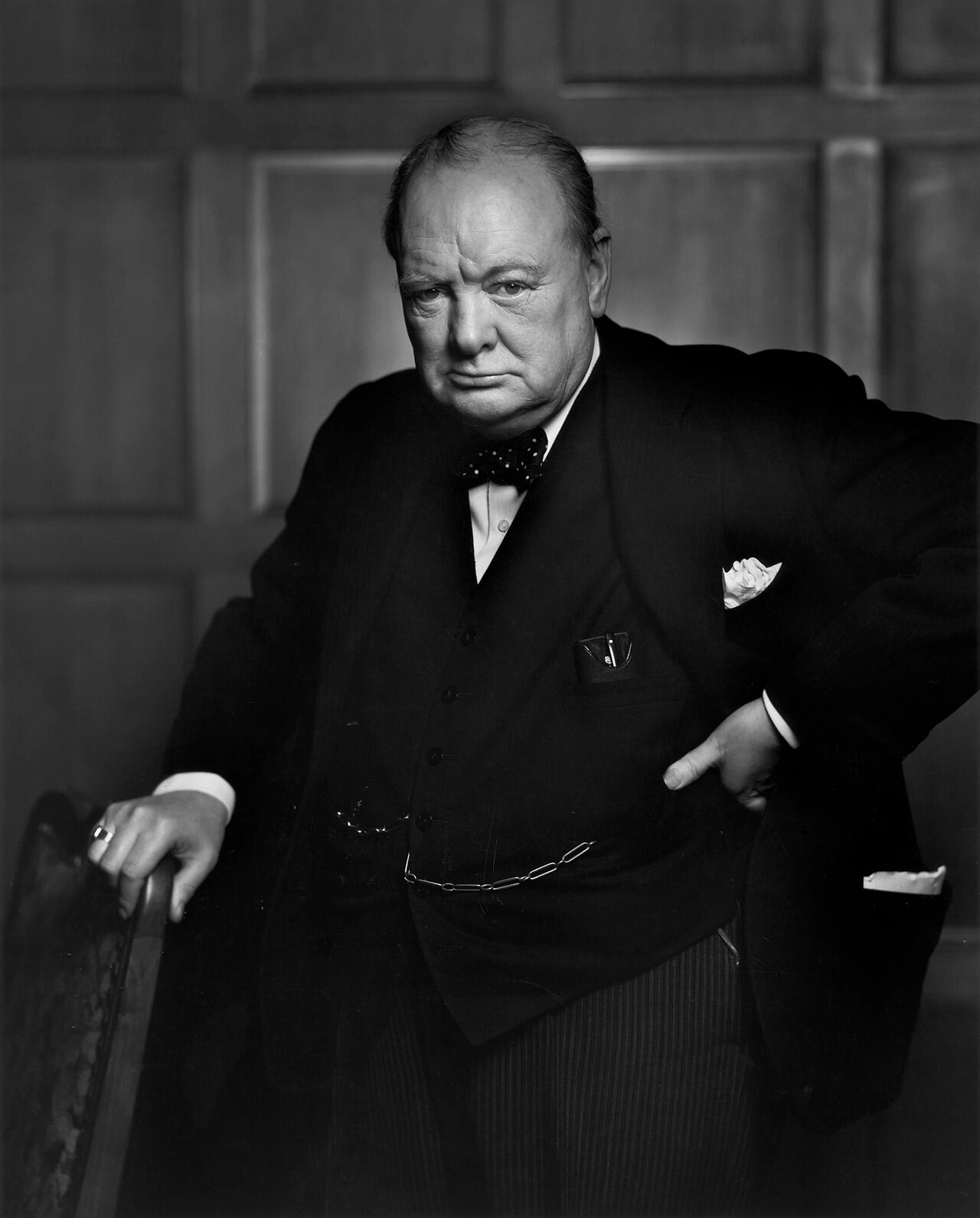 A man in a dark suit and bow tie stands with one hand on a chair and the other at his waist, looking sternly at the camera. The background is wooden paneling. Black and white photo.