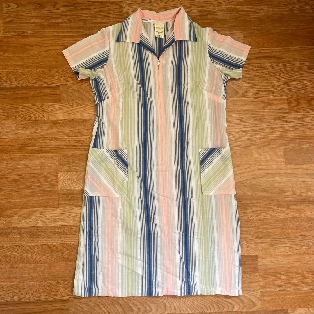 A short-sleeve, knee-length dress with vertical stripes in blue, pink, green, and white, featuring two front pockets and a collar, laid flat on a wooden floor.