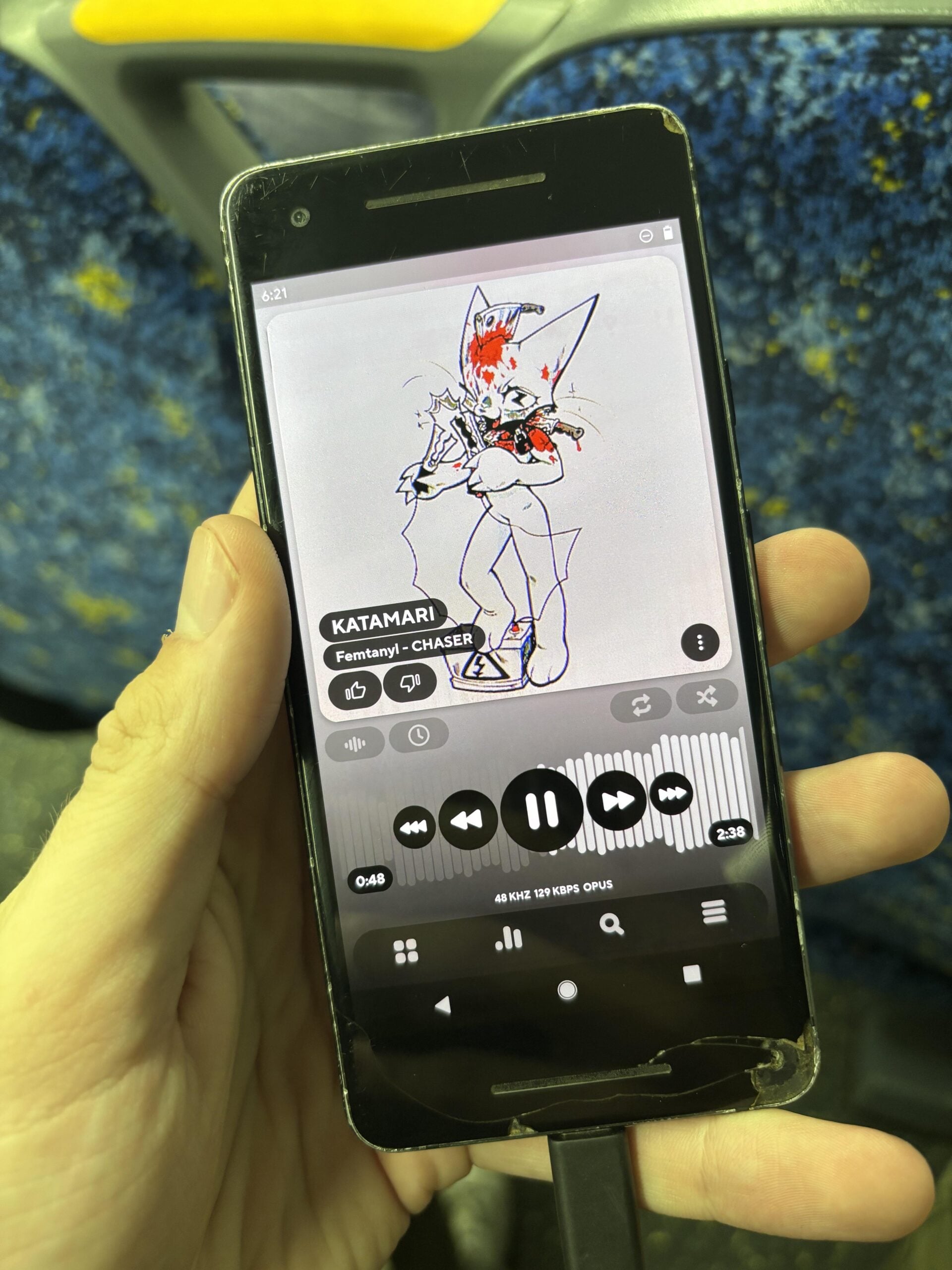 A hand holding a smartphone playing a song titled “KATAMARI” by Femtanyl + CHASER, showing digital artwork and music controls. The background is a blue patterned bus seat.