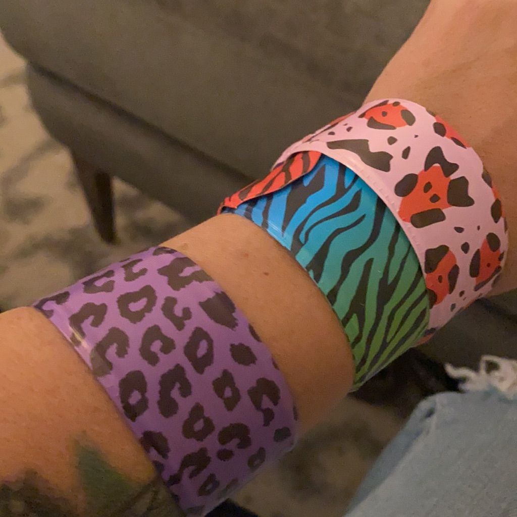 A forearm with three slap bracelets: purple leopard print, blue and green zebra stripes, and pink with red and black spots. The background is a gray couch and carpet.