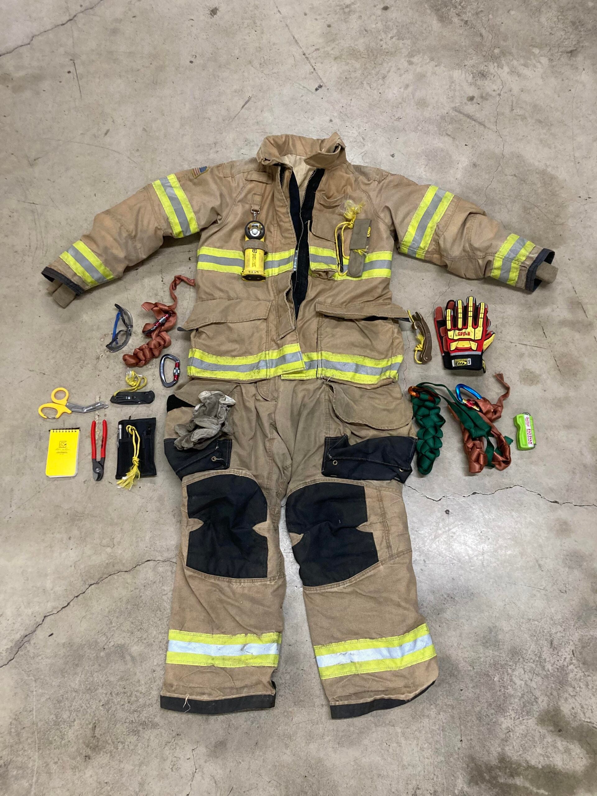 A tan firefighter suit with yellow reflective stripes is laid out on a concrete floor, surrounded by gloves, goggles, ropes, scissors, notepad, and other rescue tools.