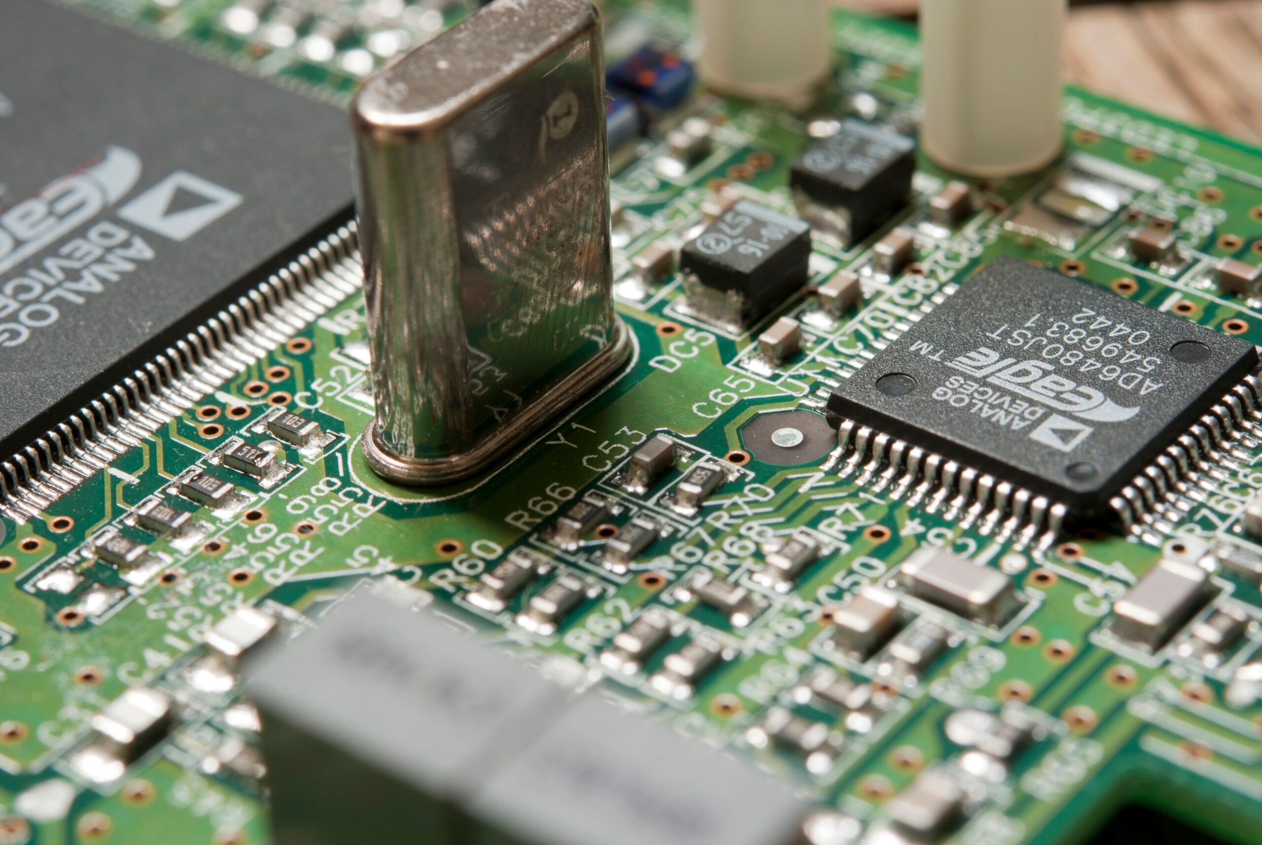 Close-up of a green circuit board showing various electronic components, including microchips, capacitors, resistors, and a metal crystal oscillator.