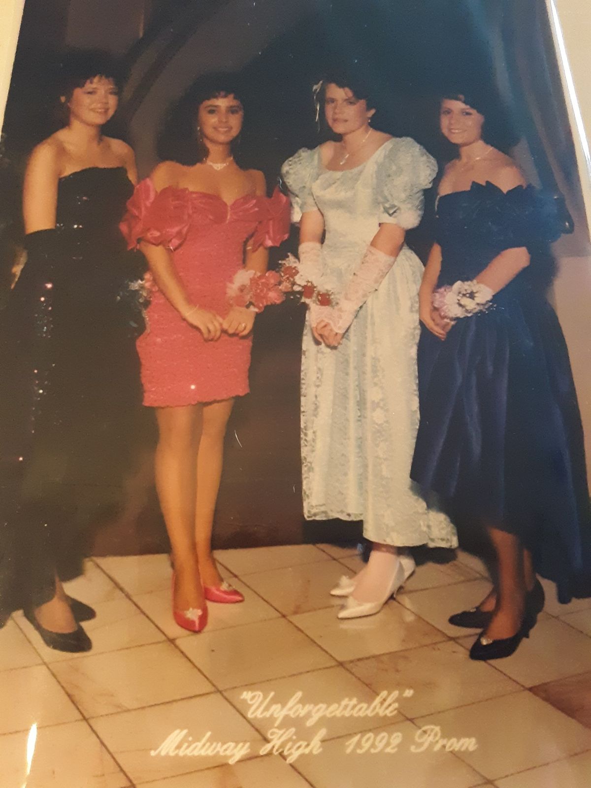 Four young women in prom dresses stand together at the “Unforgettable” Midway High 1992 Prom. They wear corsages and pose indoors, smiling at the camera. Two wear dark dresses, one wears red, and one wears light blue.