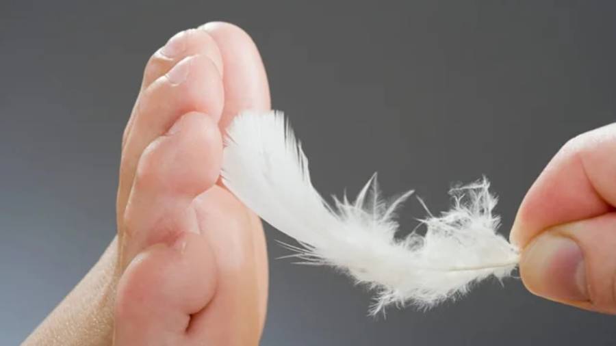A close-up of a person’s bare foot being lightly touched by a white feather, suggesting ticklishness or sensitivity, with a plain gray background.