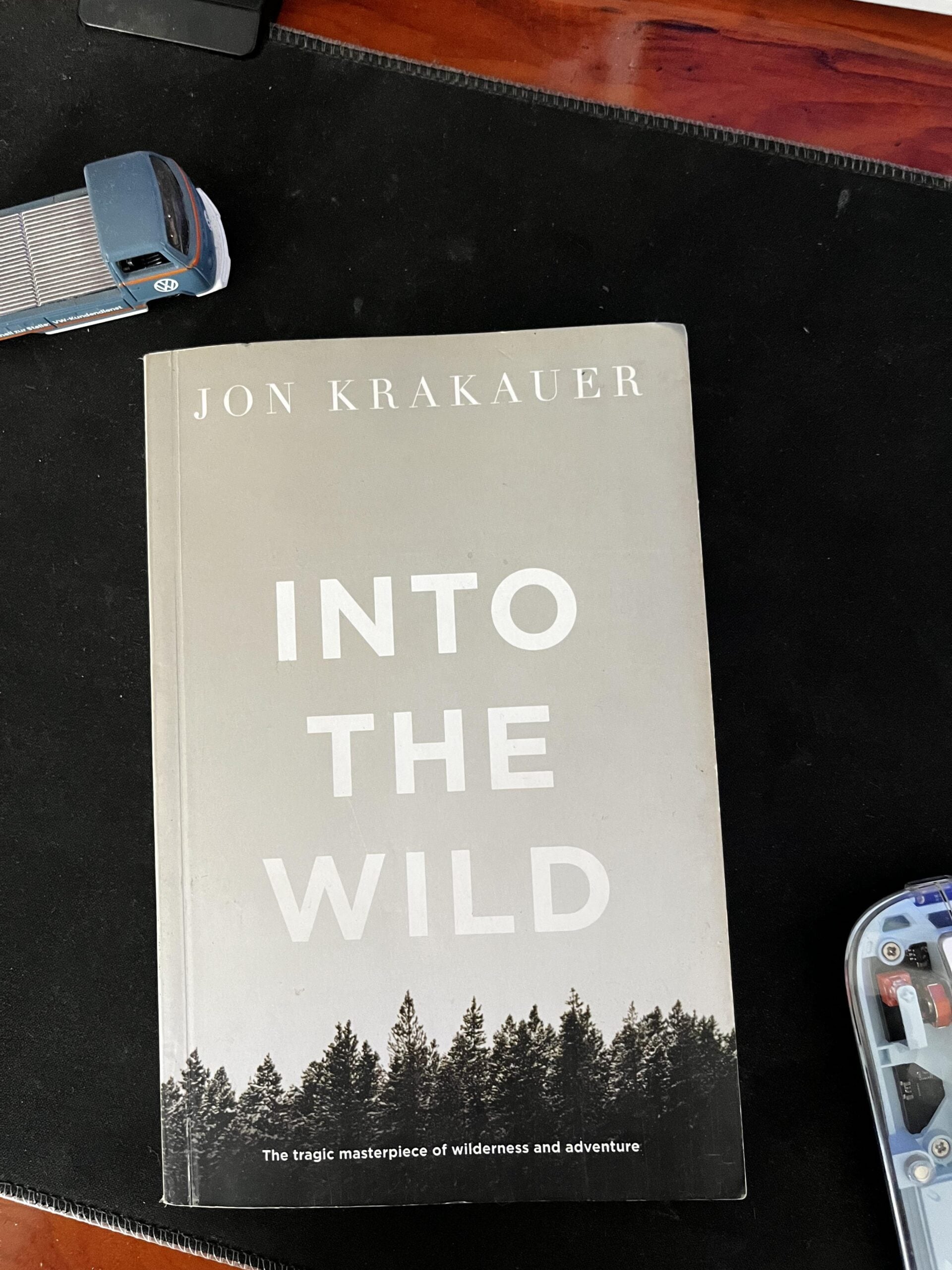A paperback copy of “Into the Wild” by Jon Krakauer lies on a black surface next to a blue toy car and a clear plastic case.