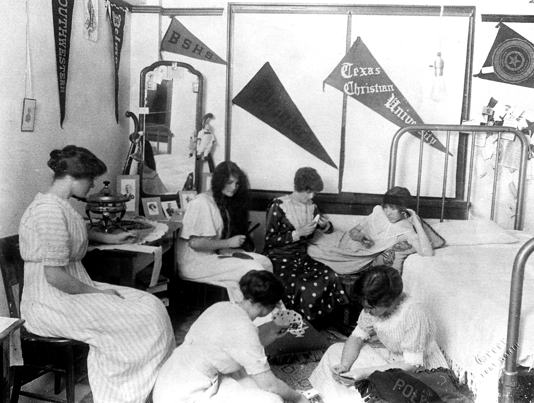 Five young women sit and sew in a dorm room decorated with pennants, a mirror, and a bed. The setting appears to be a college residence, with vintage clothing and furnishings.