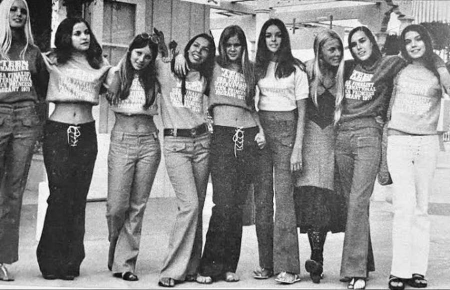 A group of eight young women in 1970s fashion—crop tops, flared pants, and long hair—stand arm in arm outdoors, smiling at the camera.