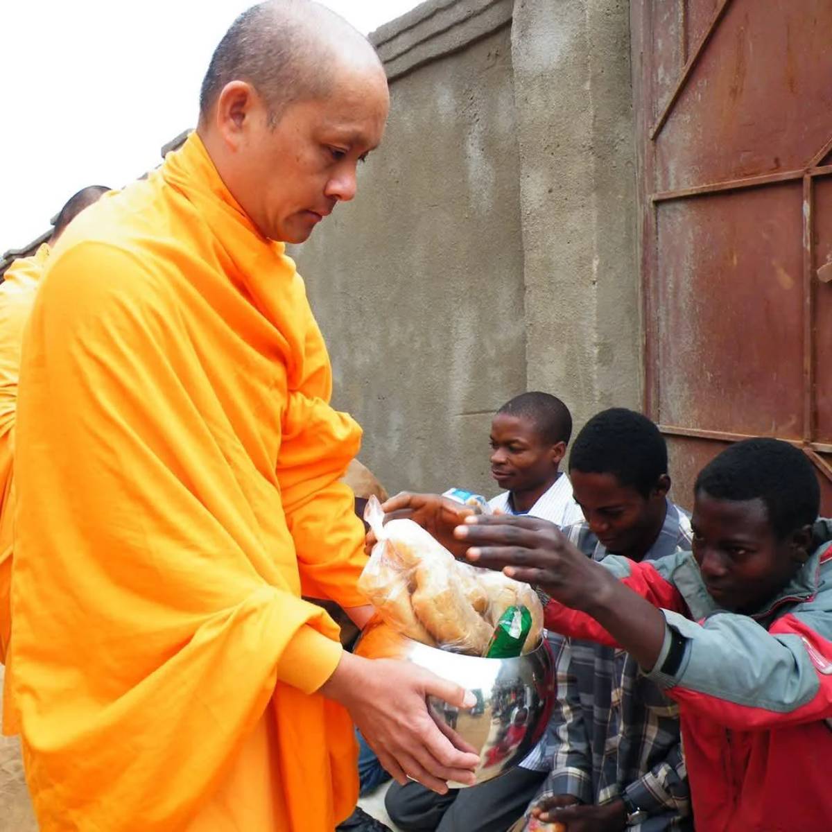 A Buddhist monk in an orange robe receives food offerings from seated men, placing items into a silver bowl, against a concrete wall and metal gate.