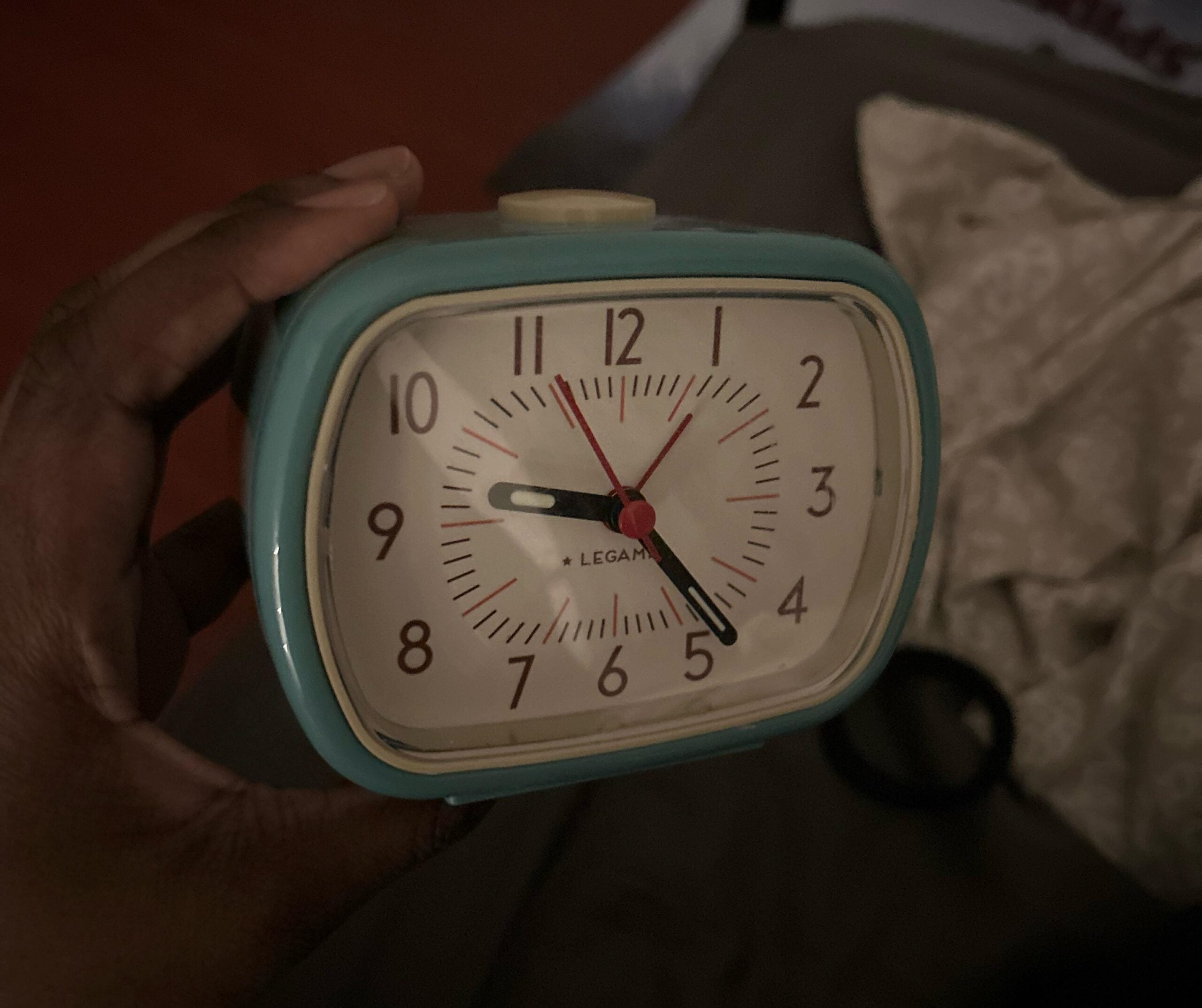 A hand holding a small, rectangular blue alarm clock showing 7:42. The background includes a patterned cloth and a dimly lit room.