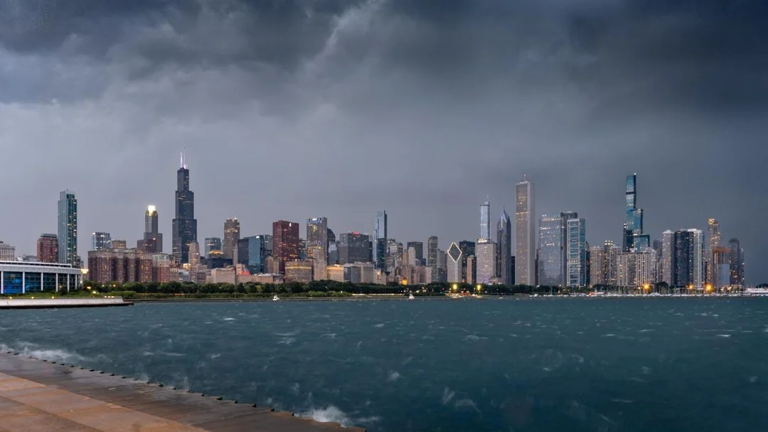 Chicago skyline with tall buildings under a dark, cloudy sky, viewed across Lake Michigan from a waterfront promenade.
