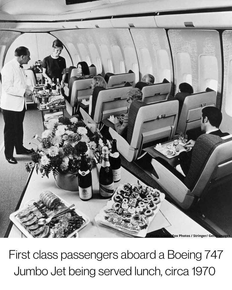 A black and white photo of first-class passengers on a Boeing 747 in 1970, seated at tables as flight attendants serve an elaborate lunch with flowers, wine, and various dishes.