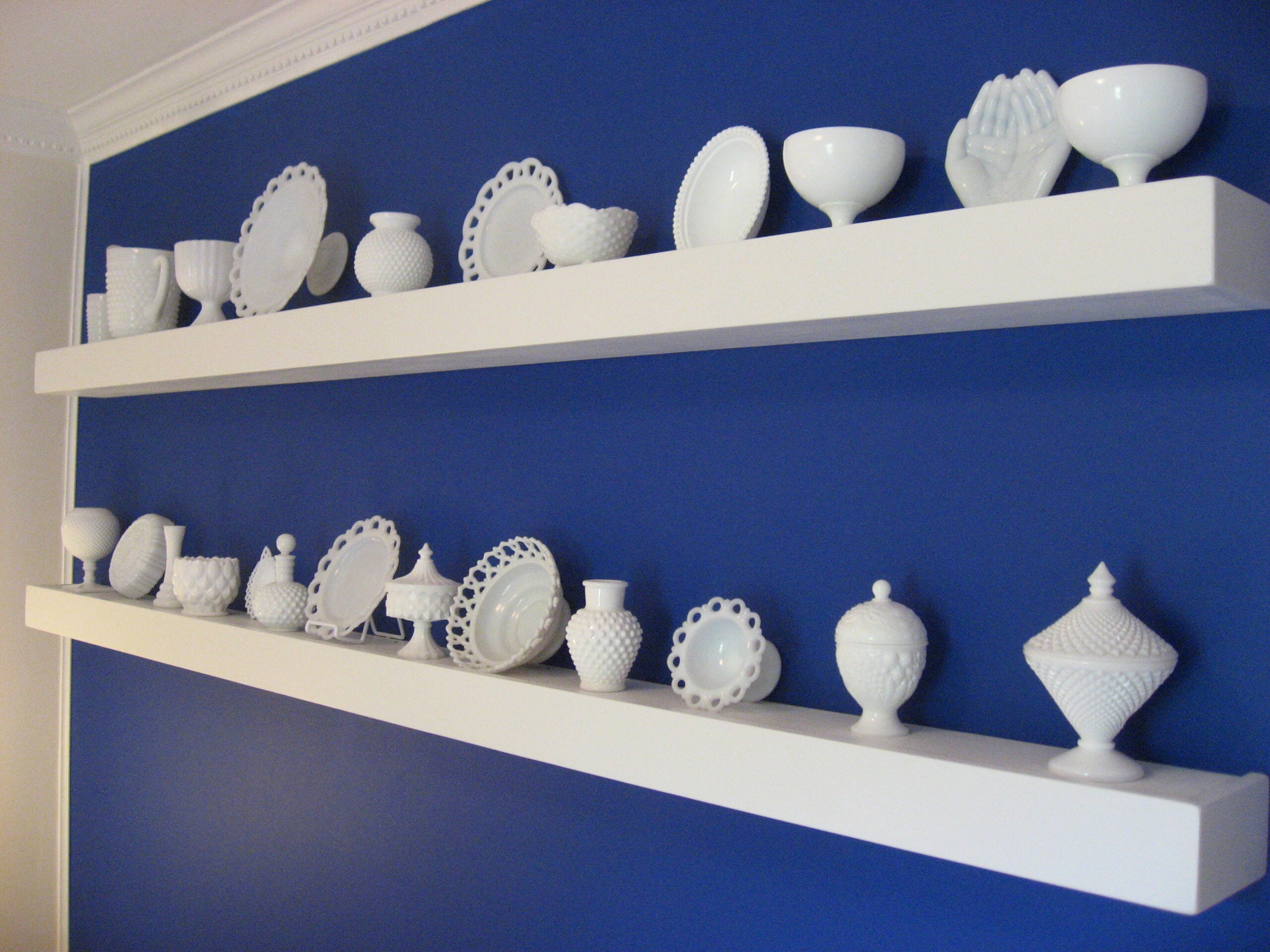 Two white floating shelves on a blue wall hold various white ceramic dishware, including plates, bowls, vases, and decorative pieces, neatly arranged in rows.