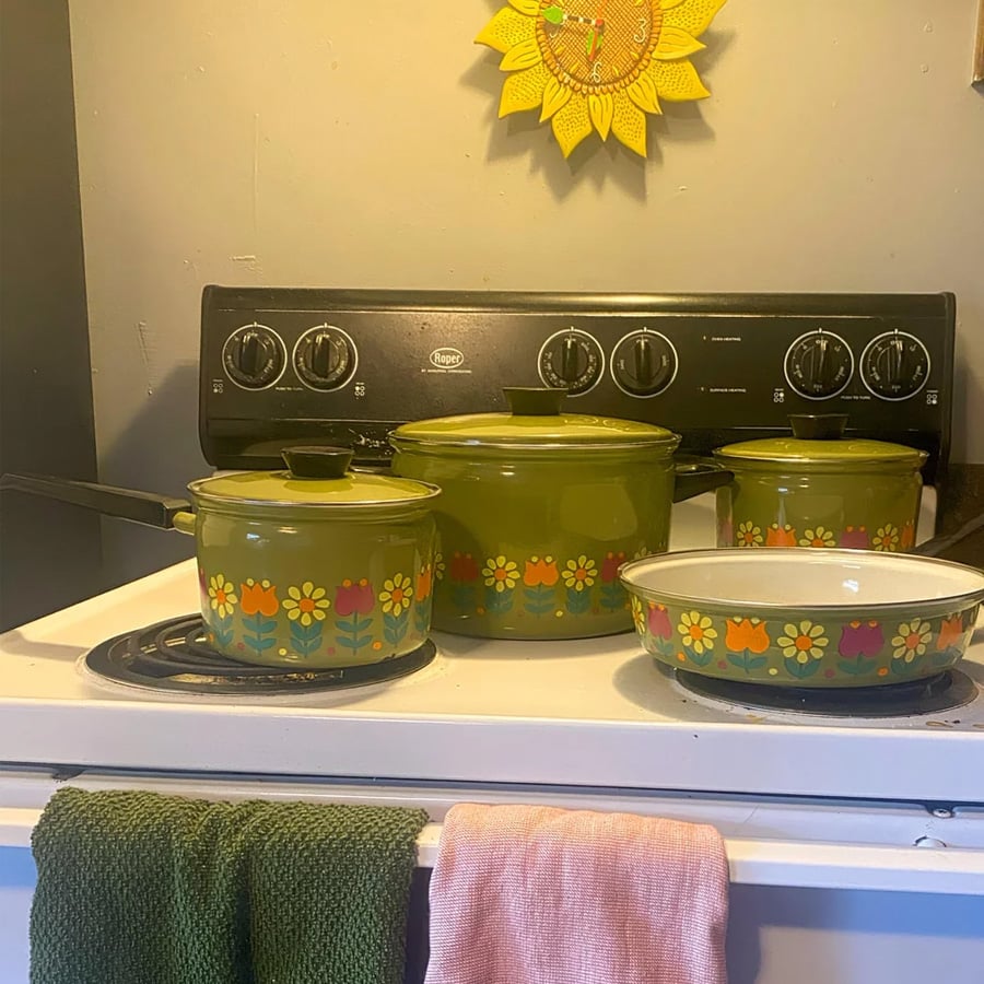 Four vintage green pots with flower patterns sit on a stove. Two towels hang from the oven handle, one green and one pink. A yellow sun decoration hangs on the gray wall above the stove.