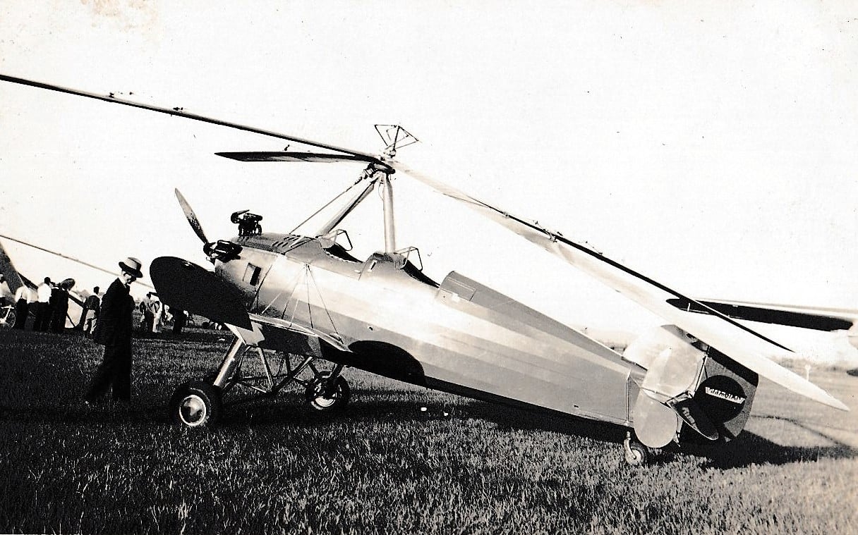 A vintage autogyro aircraft sits on grass, with a person in a hat walking nearby and a small crowd gathered in the background.