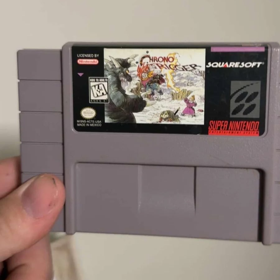 A hand holds a Super Nintendo “Chrono Trigger” game cartridge. The label features fantasy artwork and logos for Nintendo, Squaresoft, and the Super Nintendo Entertainment System.