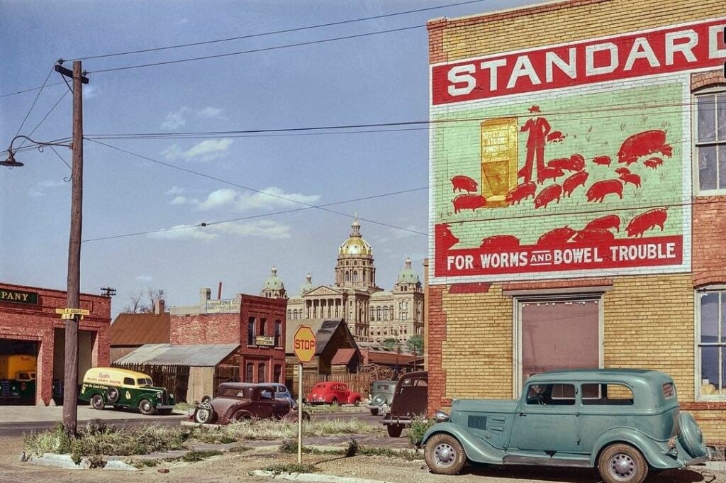 A vintage street scene with old cars, brick buildings, a large “Standard” ad on a wall, and a domed government building in the background under a blue sky.