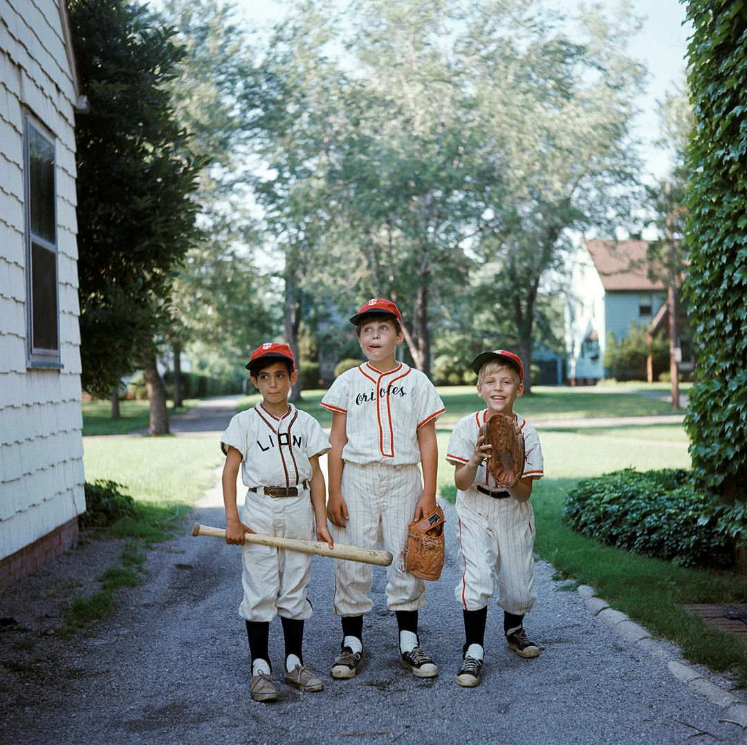 Three young boys in vintage baseball uniforms and caps stand outside on a path; one holds a bat, one a mitt, and one a glove. Trees and houses are visible in the sunny background.