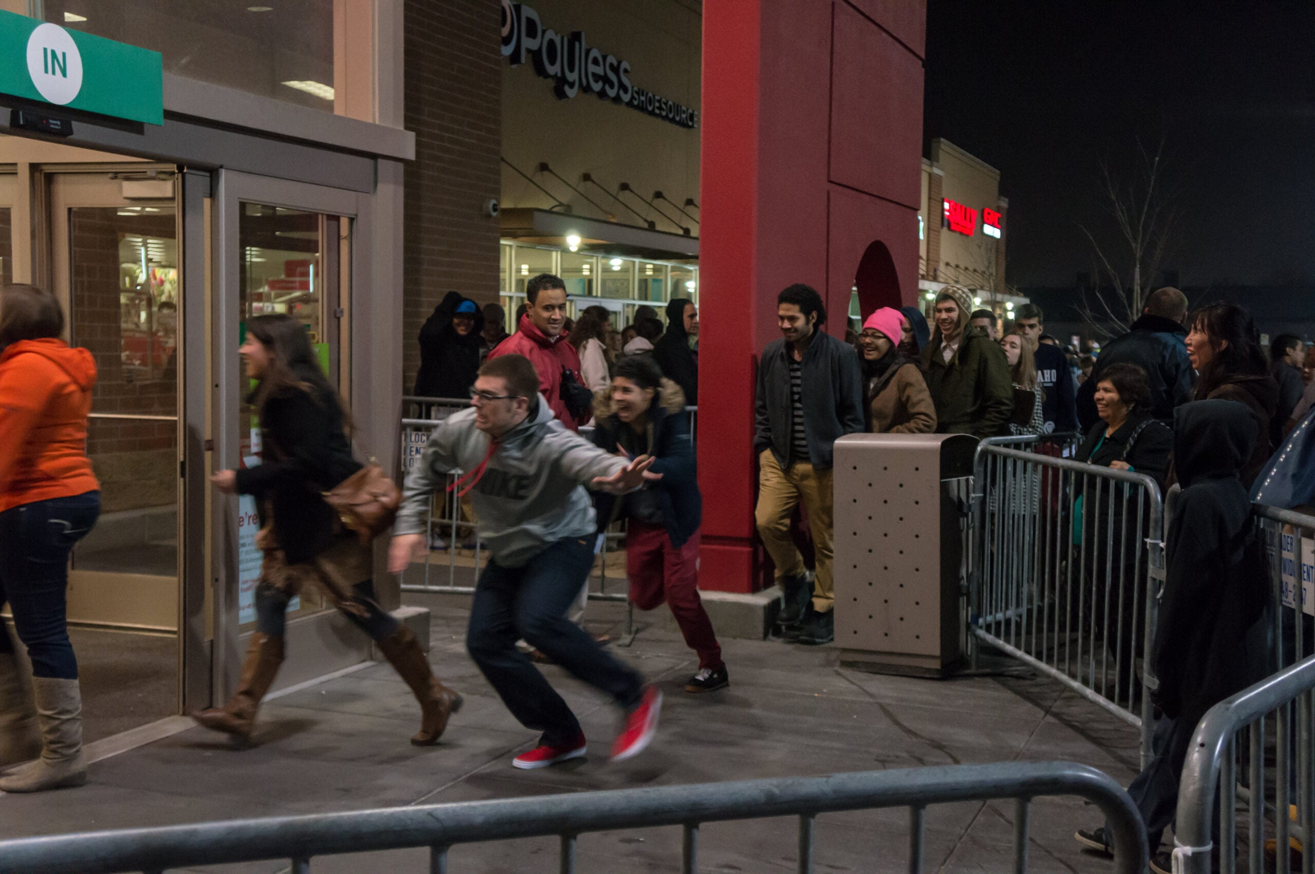 Shoppers rush through store doors at night as a crowd waits outside behind barriers, likely for a big sale or event.
