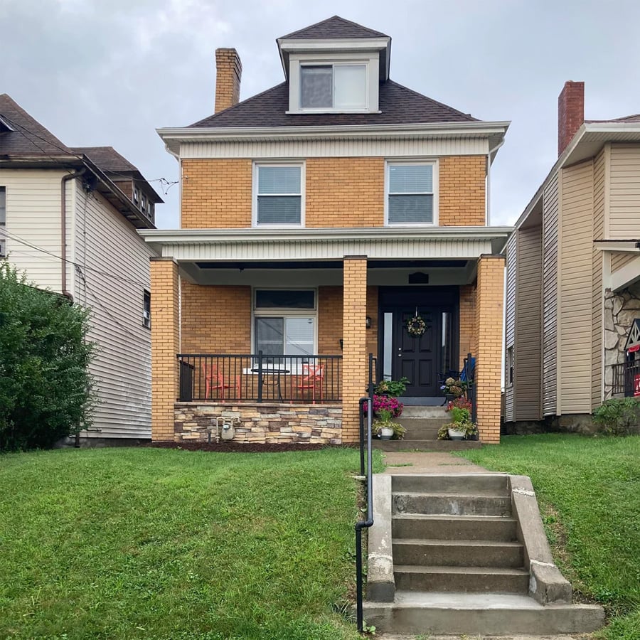 A two-story brick house with a front porch, potted plants by the steps, a dark front door with a wreath, and a well-kept green lawn, situated between two neighboring houses on a cloudy day.