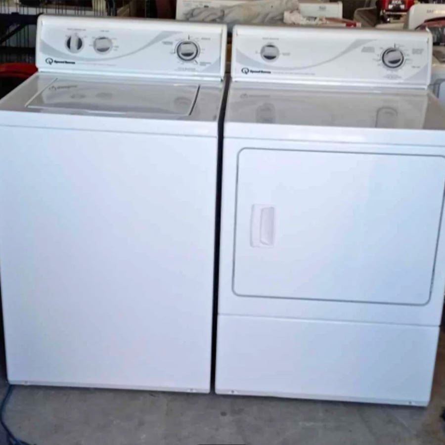 A white top-loading washing machine sits next to a matching front-loading dryer. Both appliances have control knobs and are placed side by side on a concrete floor in what appears to be a laundry or storage area.