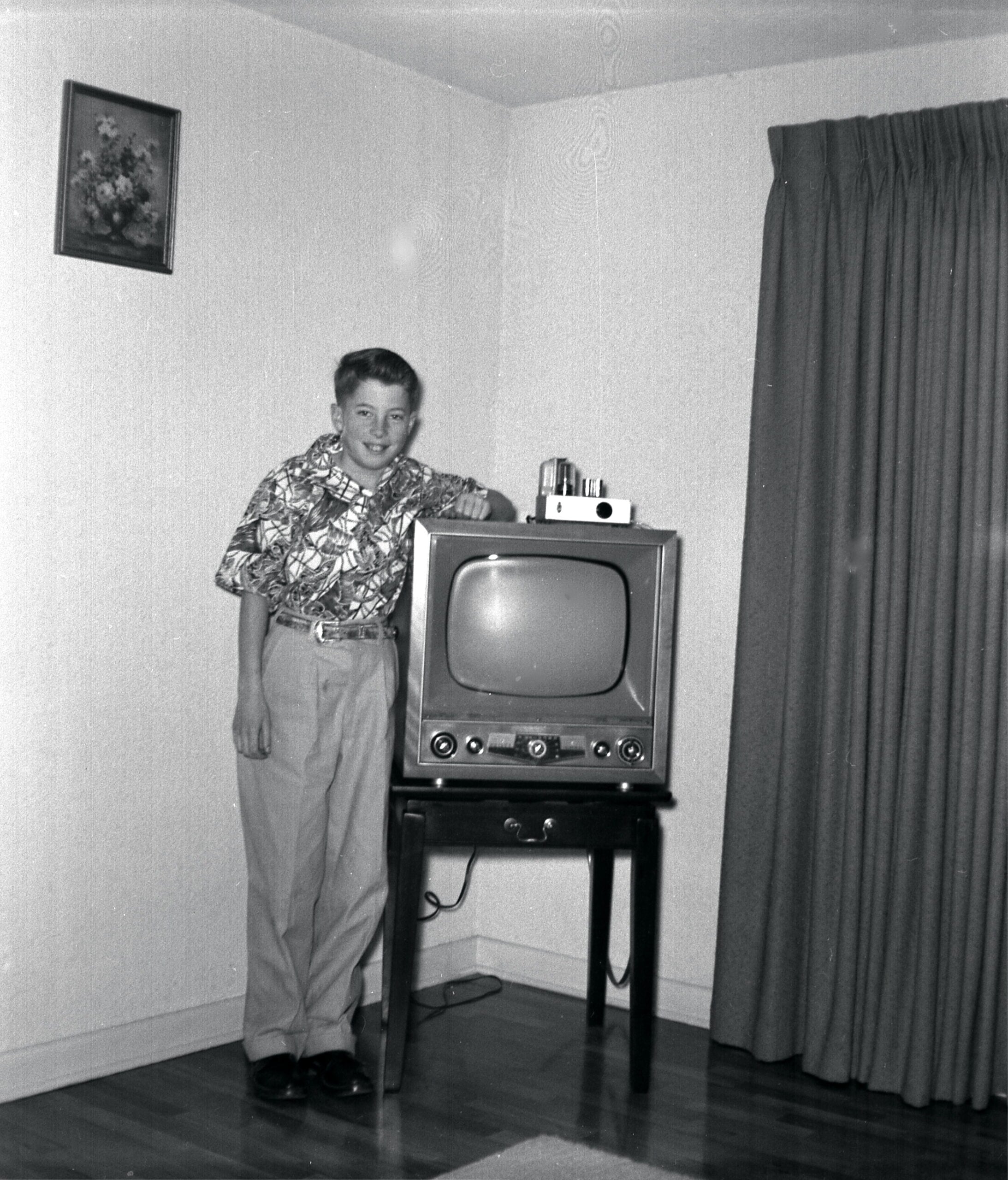 A boy in a patterned shirt stands by a vintage television set on a small table in a living room, with a curtain and a framed picture on the wall behind him.