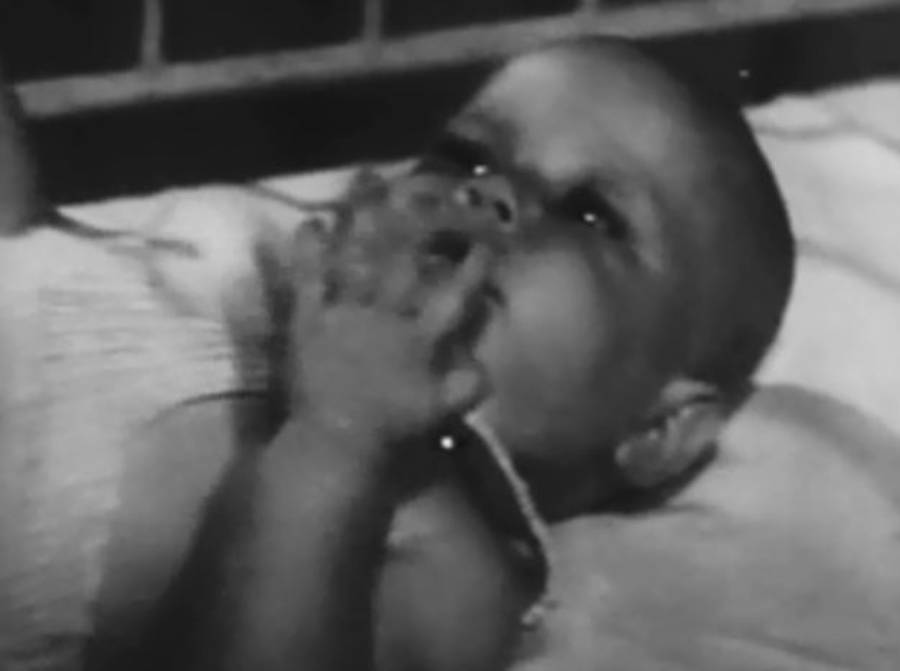 A black and white photo of a baby lying on a pillow, looking upward, with one hand resting near its mouth. The baby appears thoughtful or curious. A brick wall is visible in the background.